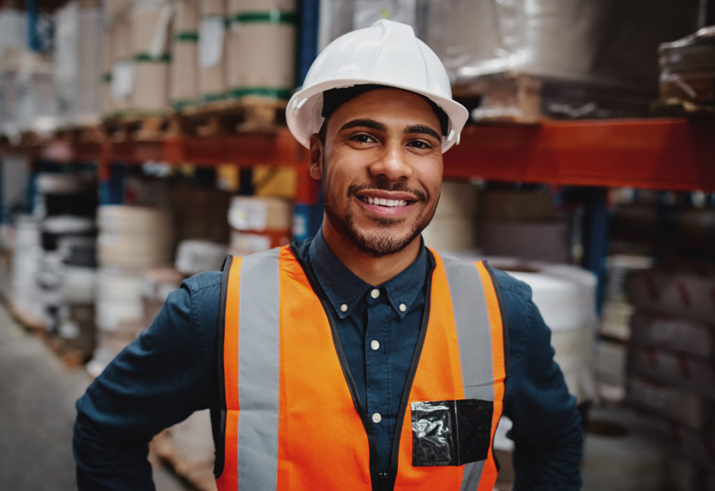 Warehouse manager smiling standing in warehouse wearing white helmet and orange safety vest